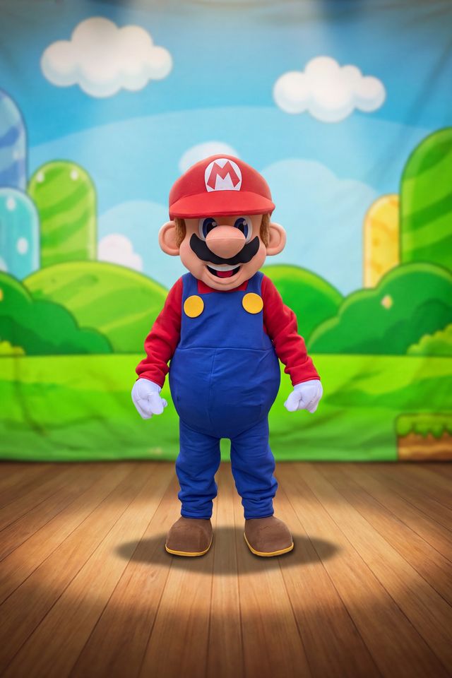 Mario en costume de mascotte, sur une scène de théâtre, devant un paysage de jeu vidéo coloré.